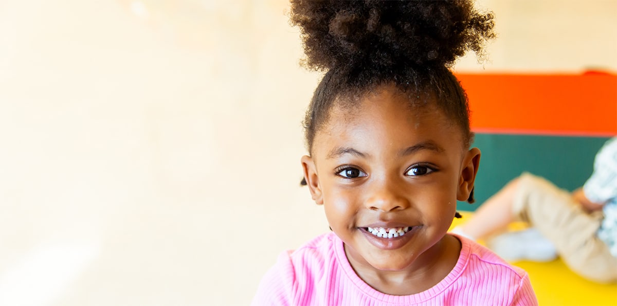 Little girl in a pink t-shirt smiling at the camera. There are bright colors in the background of red, green, and yellow.