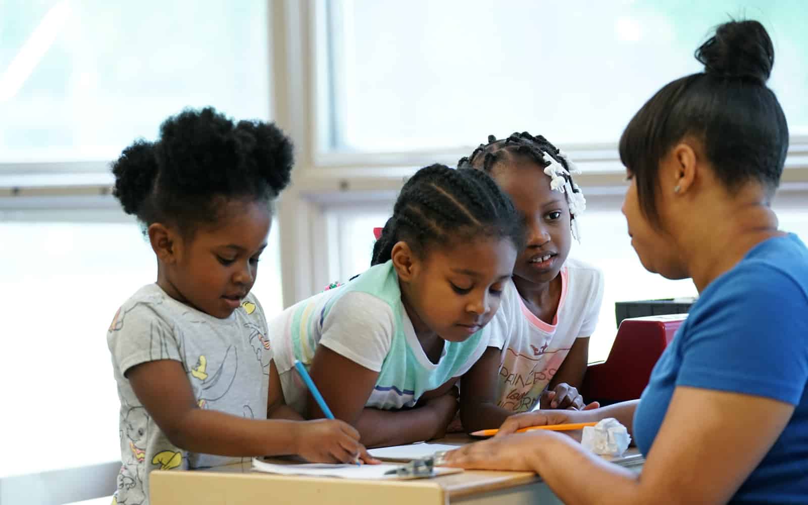 Three girls writing at table with their teacher