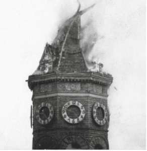 Black and white photo from 1928 of the Old Capitol Building's octagonal clocktower burning and collapsing in smoke with the hands of the clock frozen at 4:47pm.