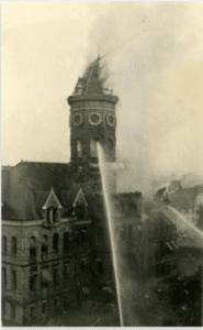 Black and white photo from 1928 of the Old Capitol Building in smoke with firefighters outside the frame spraying the building with water to put out the fire raging inside the building. One stream of water hits the right side of the building while another hits the octagonal clocktower.
