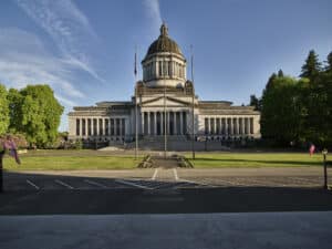 Photo of the Washington State Capitol Building in Olympia, Washington on a sunny day with three flag poles, a grassy lawn, and a parking lot at the front of the building.