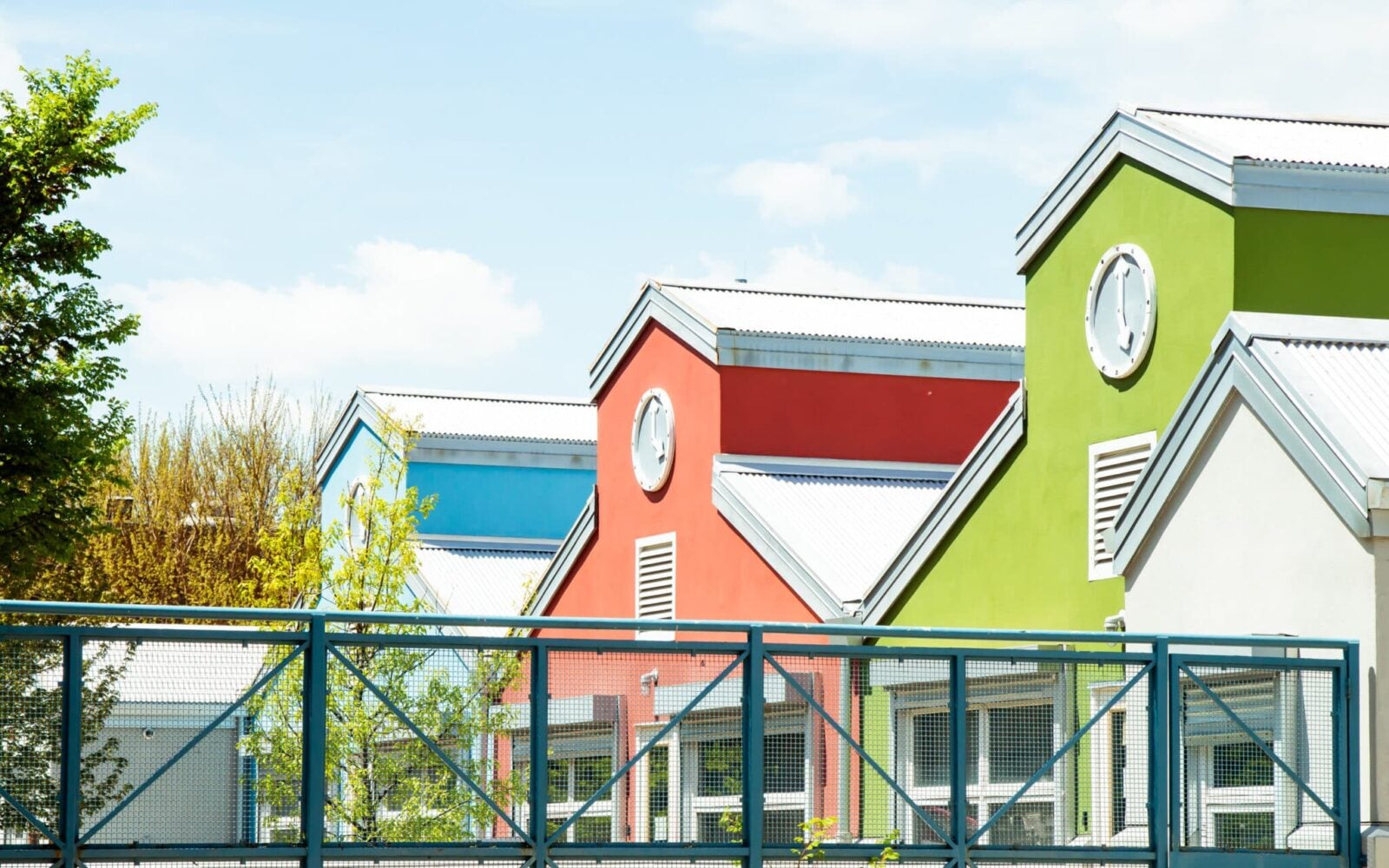 3 buildings sitting next to one another, behind a fence. The leftmost building is blue, the center building red, and the closest, rightmost building is green.
