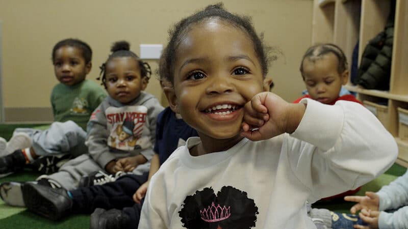 A group of young children in a classroom setting with the focus on a young boy smiling at the camera