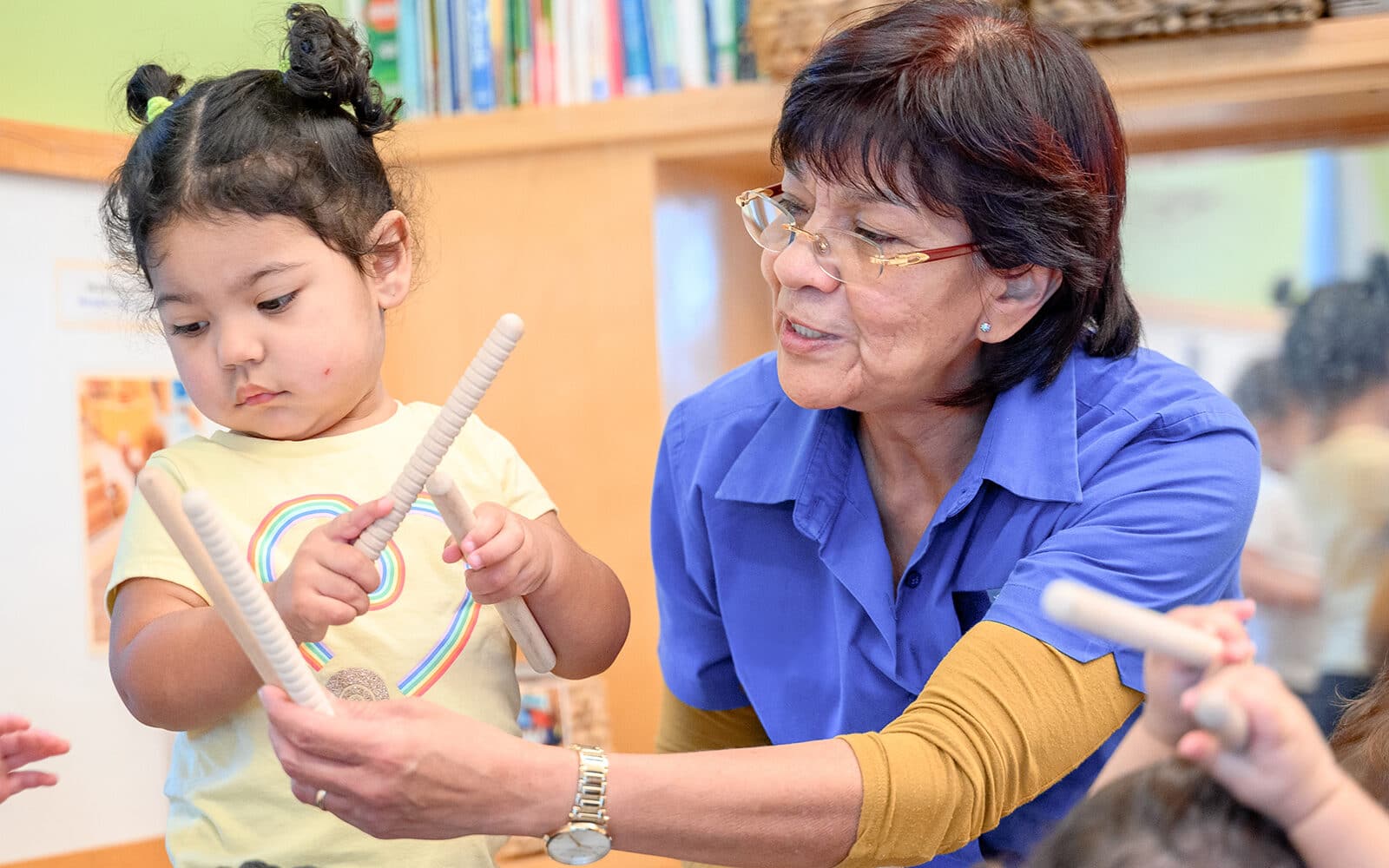 Image of child care provide in a blue polo over a yellow shirt showing drumsticks to a small child who is also holding drumsticks.