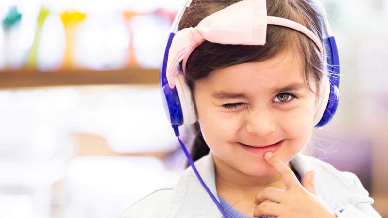 Little girl smiling with one eye closed wearing a bow and headphone set